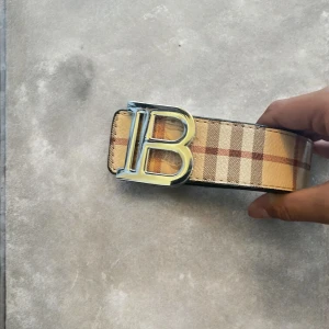 Burberry bälte med B-spänne - Två snygga Burberry-bälten med ikoniskt rutigt mönster och stort silvrigt B-spänne. Ett bälte i klassisk beige med bruna och röda rutor, det andra i svart med diskret rutmönster. Båda är breda och tillverkade i skinn, perfekta för att lyfta din outfit.