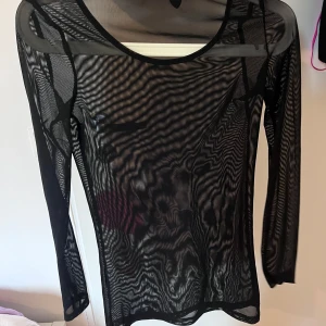 Svart mesh genomskinlig långärmad topp - Snygg svart mesh topp med lång ärm och rund hals. Tunt och transparent material som ger en edgy vibe, perfekt att styla lager på lager eller med ett coolt linne under. Passar dig som gillar att sticka ut och vill ha något trendigt i garderoben.