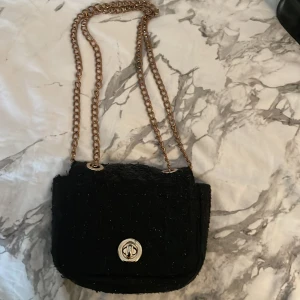 Svart axelväska med kedja - Snygg svart axelväska med guldig kedjerem och vridlås framtill. Väskan har en mjuk, lite glittrig yta och är perfekt för att lyfta din outfit. Den är liten och smidig, rymmer det viktigaste och har ett klassiskt men trendigt uttryck.