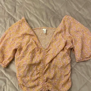 Blommig v-ringad blus från H&M - Supersöt blommig blus från H&M i ljusgult och rosa med v-ringning och rynkdetalj framtill. Blusen har korta puffärmar och smockad rygg för extra komfort. Perfekt för vår och sommar!