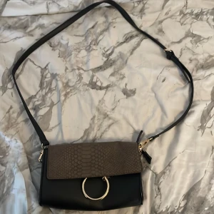 Svart axelväska med ormmönstrat lock - Snygg svart axelväska med justerbart band och lock i brunt ormmönstrat material. Väskan har guldfärgade detaljer, stor ring framtill och kedja på insidan. Perfekt för dig som vill ha en trendig och stilren accessoar med det lilla extra.