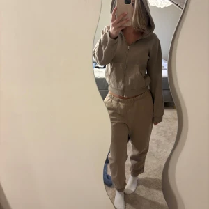 Beige mjukisbyxor med resår och Hoddie till - Säljer ett par beige mjukisbyxor med resår i midjan och muddar vid bensluten. Byxorna har en loose passform och är tillverkade i mjukt bomullstyg, perfekta för chill dagar hemma. Matchar snyggt med en hoodie eller t-shirt. Säljer även Hoddien som matchar men går att köpas enskilt 