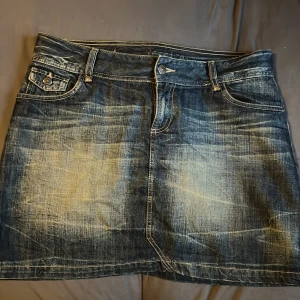 Mörkblå jeanskjol med fickor - Cool kort jeanskjol i mörkblå tvättad denim med slitna detaljer och kontrastsömmar. Kjolen har fram- och bakfickor med lock och knappar samt bälteshällor. Snygg detalj med röd broderad ruta bak. Perfekt för en avslappnad streetstil.