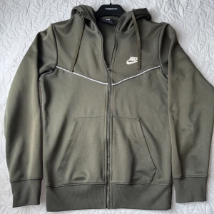 Olivgrön Nike hoodie med dragkedja XS - Nike hoodie i olivgrönt med vit logga på bröstet och snygg detaljsöm över bröstet. Tröjan har huva med snörning, två fickor fram och hel dragkedja. Perfekt för chill eller träning, storleken är xs men passade även mig som har m