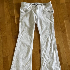 Vita lågmidjade bootcut jeans med nitar - Snygga vita lågmidjade jeans med bootcut passform. Byxorna har coola silverfärgade nitar på fickorna fram och bak samt broderad detalj vid linningen. Tillverkade i bomull med klassisk jeanskänsla och låg midja. Säljer då dom är för små för mig. Pris kan diskuteras!