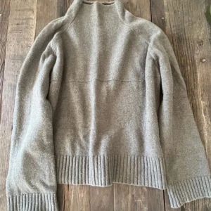 stickad tröja By Malene Birger - Mysig grå stickad tröja från By Malene Birger med hög hals och breda ribbade muddar vid ärmslut och nederkant. Tillverkad i en mjuk mix av merinoull, polyamid och bomull. Perfekt för lager-på-lager och chill höst- och vinterstil.