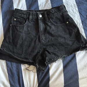 Svarta jeansshorts från Gina Tricot - Svarta jeansshorts från Gina Tricot med råa, fransiga kanter och klassisk femficksdesign. Hög midja och silverfärgad knapp framtill. Perfekta för en avslappnad och cool look under sommaren.