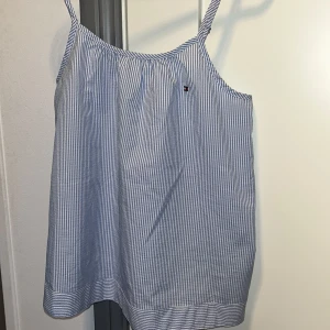 Randig linnetopp från Tommy Hilfiger - Säljer en blåvit randig linnetopp från Tommy Hilfiger med tunna axelband och klassisk logga broderad på bröstet. Toppen är luftig och har en avslappnad passform, perfekt för varma dagar. Tillverkad i mjuk bomull som känns skön mot huden.