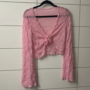 Rosa omlott blus med knyt - Säljer en superfin rosa blus med vågigt mönster och transparent tyg. Blusen har långa ärmar, croppad passform och knyt framtill. Perfekt för dig som vill ha en lätt och luftig topp med trendig look.
