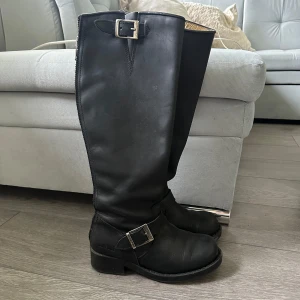 Svarta höga Johnny Bulls boots i skinn - Snygga svarta höga boots från Johnny Bulls i äkta skinn. Klassisk modell med rund tå, grov sula och två silvriga spännen. Dragkedja baktill för enkel påtagning. Perfekta för dig som gillar en tuff och stilren look. Har en liten defekt vid framsida  (kolla sista bilden)