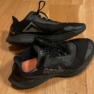 Nike Pegasus Trail Gore-Tex sneakers - Svarta Nike Pegasus Trail sneakers med Gore-Tex-material som gör dem vattentäta. Skorna har snörning, grov sula för extra grepp och diskreta reflexdetaljer. Perfekta för löpning eller promenader i alla väder. Snygg, sportig look med orange innersula och klassisk Nike-logga.