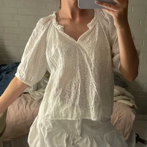 Vit blus med broderade detaljer - Superfin vit blus i bomull med broderade hålmönster och lätt puffiga, korta ärmar. Blusen har v-ringning med volangkant och en luftig, avslappnad passform. Perfekt för en fräsch och somrig look!