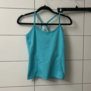 Turkos linnetopp med smala axelband  - Säljer en turkos linnetopp från Röhnisch med smala axelband som korsas i ryggen. Toppen har meshdetaljer på sidorna och baksidan för extra ventilation. Perfekt för träning eller varma dagar när du vill ha något svalt och sportigt.