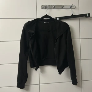 Svart croppad hoodiejacka från SHEIN - Svart croppad hoodiejacka från SHEIN med dragkedja framtill och huva. Jackan har långa ärmar och en enkel, clean look som passar perfekt till streetwear. Tillverkad i mjuk bomullsmix för en skön känsla.