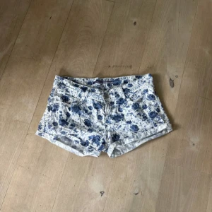 Lågmidjade shorts - Supersnygga vita shorts från Stradivarius med blått blommigt mönster. Klassisk femficksmodell med låg midja och korta ben. Perfekta för varma dagar och ger en fräsch vibe till din sommarstil. Låg midjade tyg shorts