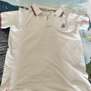 Vit Moncler pikétröja med rödblå detaljer - Snygg vit pikétröja från Moncler med klassisk krage och knappar framtill. Tröjan har Moncler-logga på bröstet och coola rödblå ränder på krage och ärmslut. Tillverkad i mjuk bomull som känns skön mot huden. Perfekt för en clean och sportig look.