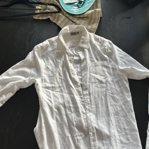 Vit linneskjorta från RYVLS - Klassisk vit skjorta från RYVLS i svalt linne. Skjortan har lång ärm, bröstficka och knappar framtill. Perfekt för en clean och stilren look, lätt att matcha med jeans eller shorts. Snygg krage och avslappnad passform.