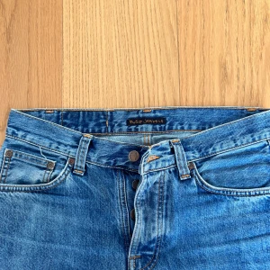 Blå jeans från Nudie Jeans Co - Snygga blå jeans från Nudie Jeans Co med klassisk femficksdesign och orange sömmar. Byxorna har raka ben, hög midja och ikonisk broderad detalj på bakfickorna. Tillverkade i slitstarkt denim med knappgylf och skön passform.
