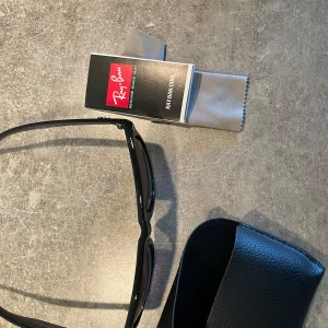 Svarta Ray-Ban solglasögon - Klassiska svarta Ray-Ban solglasögon med glansig båge och mörka glas. Säljes med originalfodral, putsduk och Ray-Ban booklet. Snygg och tidlös modell som ger en cool vibe till din stil.