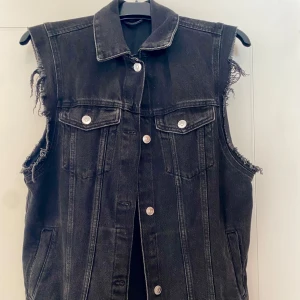 Svart jeansväst med råa kanter - Cool svart jeansväst med råa, fransiga kanter vid ärmhålen och klassisk krage. Västen har två bröstfickor med lock och silverfärgade knappar framtill. Perfekt att slänga över en hoodie eller t-shirt för en edgy look. På etiketten står det att den är xs men passar män med storleken M 