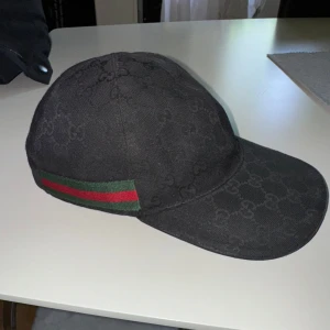 Gucci keps - Snygg svart keps från Gucci med diskret GG-logomönster över hela kepsen. Klassisk böjd skärm och ikoniska gröna och röda ränder på sidan. Tillverkad i Italien och i ett slitstarkt bomullsmaterial. Perfekt för dig som vill ha en exklusiv streetstyle-look. Storlek S, äkta såklart.