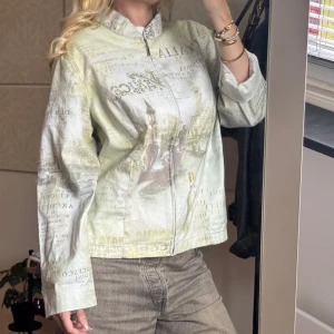 Overshirt med vintage print och dragkedja - Säljer en oversized overshirt i ljusgrön och beige ton med unikt vintageinspirerat print och textdetaljer. Jackan har dragkedja framtill, ståkrage och långa ärmar. Perfekt för dig som gillar statement-plagg med konstnärlig vibe.