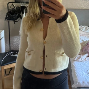 Kofta med svarta detaljer - Snygg kofta med svarta detaljer vid ärmslut och nederkant. Guldiga knappar framtill och två små fickor med dekorativa knappar. Passar perfekt till highwaist jeans eller kjol. Köpt på H&M och aldrig använd i storlek S🤍