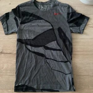 Nike Dri-Fit grå t-shirt med mönster - Nike Dri-Fit t-shirt i grått med svart abstrakt mönster och röd swoosh-logga på bröstet. Klassisk rund hals och korta ärmar. Perfekt för dig som gillar sportig och stilren look.