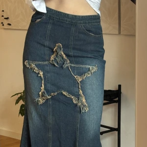 Jeanskjol med stjärndetalj och fransar - Unik lång jeanskjol i blå denim med stor fransad stjärna framtill och rå fransad nederkant. Kjolen har hög midja, dragkedja i sidan och klassiska jeansfickor. Perfekt för dig som vill sticka ut med en edgy och kreativ look.