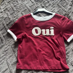 Röd t-shirt med Oui-tryck - Cool röd t-shirt med vit text 'Oui' framtill. T-shirten har vita detaljer vid halsringning och ärmslut. Perfekt för dig som gillar franska vibbar och vill sticka ut lite extra. Skön passform och mjukt bomullsmaterial och jah köpte den från spanien den är skit fin.