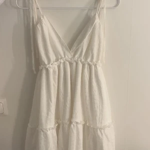 Vit omlottklänning med volanger - Superfin vit kortklänning med omlott och volangdetaljer. Klänningen har smala axelband som knyts, v-ringning och en luftig, rynkad kjol. Perfekt för varma dagar och har en romantisk vibe.