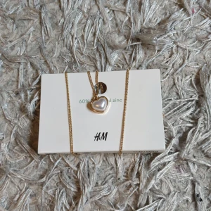 Halsband - Snyggt guldigt halsband från H&M med ett hjärtformat hänge som har en pärlvit insida. Kedjan är tunn och elegant, perfekt för att ge en söt touch till din outfit. Hjärtat ger en romantisk vibe och halsbandet är lätt att matcha med andra smycken.
