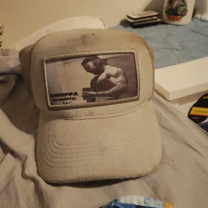 Beige meshkeps med tryck Chrippa - Säljer en beige keps med mesh på sidorna och baksidan samt justerbar snapback. Framtill finns ett svartvitt tryck av en muskulös man och texten 'CHRIPPA DA KRÄFTA!'. Perfekt för dig som gillar unika accessoarer med attityd.