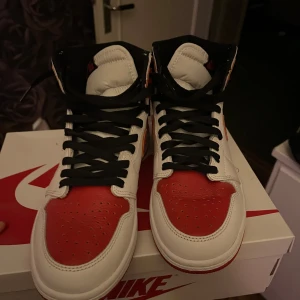 Nike Air Jordan 1 Retro High OG röd/vit - Nike Air Jordan 1 Retro High OG i klassisk röd och vit färg med svarta detaljer. Skorna har svart snörning, röd swoosh och hög siluett. Tillverkade i skinn med perforerad tå och ikonisk Air Jordan-logga på sidan. Perfekta för dig som vill sticka ut med en tidlös sneaker.