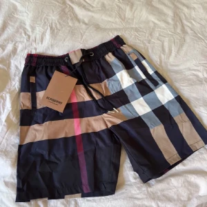 Burberry rutiga shorts med snörning - Snygga shorts från Burberry med klassiskt rutigt mönster i svart, beige, vitt och rött. De har elastisk midja med snörning och sidofickor. Perfekta för sommaren och ger en lyxig vibe till din outfit.