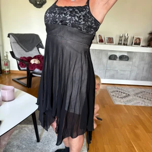 Svart kortklänning med spetsdetalj - Supersnygg svart kortklänning med transparent, flowy kjol och spets upptill. Axelbandsmodell med asymmetrisk nederkant och blommig spets över bysten. Perfekt för dig som vill sticka ut med en unik och feminin look.