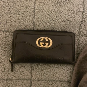 Svart plånbok från Gucci - Snygg svart plånbok från Gucci med det klassiska GG-mönstret och en elegant guldfärgad logotyp på framsidan. Plånboken har flera fack och en dragkedja för säker förvaring av dina kort och kontanter. Lite sliten då den är köpt för länge sedan men väldigt fin endå kanske för dig som gillar vintage saker🤗