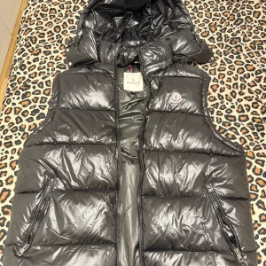 Svart dunväst från Moncler - Säljer en svart, glansig dunväst från Moncler med hög krage och två dragkedjefickor. Västen har klassisk quiltad design och ikonisk Moncler-logga på bröstet. Perfekt för lager-på-lager och streetstyle. Innerfoder med serietidningsmönster för extra cool detalj. Storlek XL