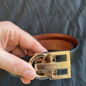 Svart och brun skärp från Salvatore Ferragamo - Stilrent vändbart bälte från Salvatore Ferragamo i svart och brun skinn med exklusivt guldspänne. Bältet har en klassisk design med präglad yta och ikoniskt metallspänne med märkets logga. Perfekt accessoar för att lyfta din outfit.