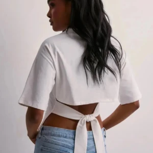 Nelly knyt topp - Säljer en vit croppad topp med korta ärmar och snygg öppen rygg som knyts baktill. Perfekt till jeans eller kjol för en trendig look. T-shirten har en enkel design och är gjord i mjukt bomullstyg.