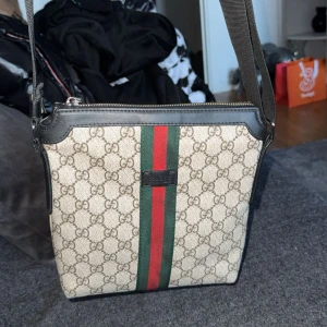 Gucci axelväska med GG-mönster - Snygg axelväska från Gucci med klassiskt GG-mönster i beige och brunt. Väskan har svarta skinn-detaljer, dragkedja upptill och den ikoniska gröna och röda randen framtill. Justerbar axelrem i textil och logopatch på framsidan.