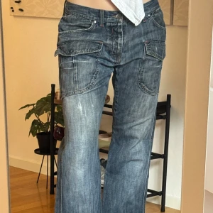 Baggy cargojeans från Benetton - Baggy blå cargojeans från Benetton med stora fickor och coola röda detaljer. Jeansen har låg midja, bred passform och är tillverkade i slitstarkt denim. Insidan av fickorna är fodrade i vinrött tyg för en extra touch.