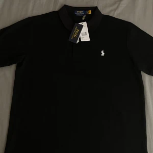 Svart pikétröja från Polo Ralph Lauren - Klassisk svart pikétröja från Polo Ralph Lauren med korta ärmar och vit broderad logga på bröstet. Tröjan har krage och knappar framtill, perfekt för en stilren och avslappnad look.