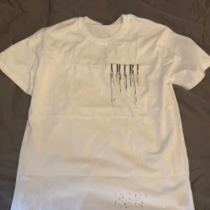Vit AMIRI t-shirt med färgstänk - Snygg vit t-shirt från AMIRI med stort tryck på ryggen och mindre tryck på bröstet. Trycket består av AMIRI-loggan med färgglada droppar i blått, orange och svart som rinner ner. Klassisk passform och rund hals, perfekt för en trendig streetwear-look.