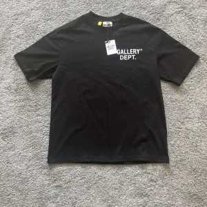 Svart t-shirt från Gallery Dept. - Svart t-shirt från Gallery Dept. med vit logga tryckt på bröstet och stor text på ryggen. Klassisk passform med rund hals och korta ärmar. Tillverkad i mjuk bomull, perfekt för en clean och stilren streetlook.