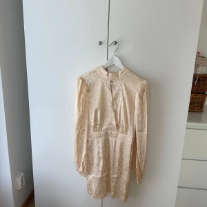 Champagnefärgad jacquardklänning NA-KD - Superfin kort klänning från NA-KD i champagnefärgad jacquard med subtilt blommönster. Klänningen har lång ärm, hög krage och en snygg slits framtill. Materialet är glansigt och känns lyxigt, perfekt för dig som vill sticka ut.