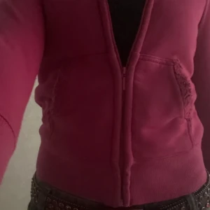 Mörkrosa zip hoodie med fransade fickor - Säljer en mörkrosa hoodie med dragkedja och fransade detaljer vid fickorna. Jackan har långa ärmar, perfekt för dig som gillar en chill och edgy stil. Materialet är mjukt och skönt, passar till jeans eller kjol. Luddigt inuti