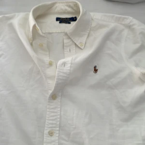 Vit skjorta från Polo Ralph Lauren m i barn passar xs - Klassisk vit skjorta från Polo Ralph Lauren i custom fit. Skjortan har knappar framtill, button-down krage och den ikoniska broderade loggan på bröstet. Perfekt för en clean och stilren look.