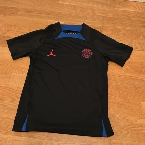 Svart PSG x Jordan fotbollströja M barn 137-147 - Svart Paris Saint-Germain fotbollströja i samarbete med Jordan. Slim fit-modell med blå detaljer vid axlar och sidor, röd broderad Jumpman-logga och PSG-märke på bröstet. Tillverkad i lätt och ventilerande material, perfekt för träning eller match.
