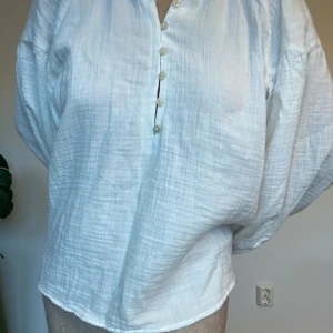 Vit henley-skjorta i bomull - Luftig och oversized vit henley-skjorta med lång ärm och knappar framtill. Skjortan är tillverkad i mjuk, strukturerad bomull och har en avslappnad passform med lätt rynkade ärmar. Perfekt för en chill och stilren look.